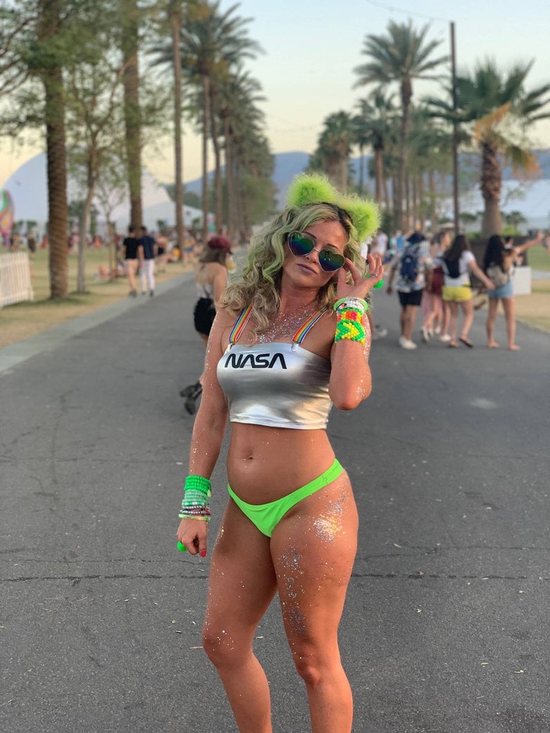 May include: A woman wearing a silver crop top with the word "NASA" printed on it, green glitter on her body, and a green bikini bottom. She is wearing sunglasses and green furry cat ears.