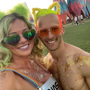 May include: A couple wearing colorful costumes and glitter at a festival. The woman is wearing a silver top with rainbow suspenders and a green wig with feather earmuffs. The man is wearing a neon yellow wig with yellow earmuffs and has gold glitter on his chest. They are both wearing sunglasses.