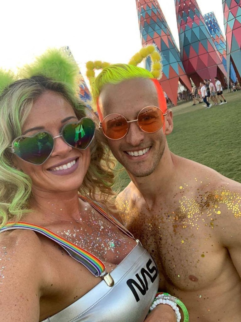 May include: A couple wearing colorful costumes and glitter at a festival. The woman is wearing a silver top with rainbow suspenders and a green wig with feather earmuffs. The man is wearing a neon yellow wig with yellow earmuffs and has gold glitter on his chest. They are both wearing sunglasses.