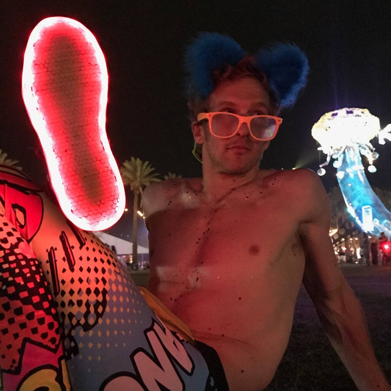 May include: A person wearing a pair of blue fuzzy bunny ears, yellow-tinted glasses, and colorful leggings with a "BOOM" graphic. The person is sitting on the ground with a red light illuminating their bare chest and the sole of a shoe.