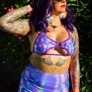May include: A woman wearing a purple iridescent halter top with a ring detail and a matching skirt. She has purple hair, a star hair clip, and large feather earrings.