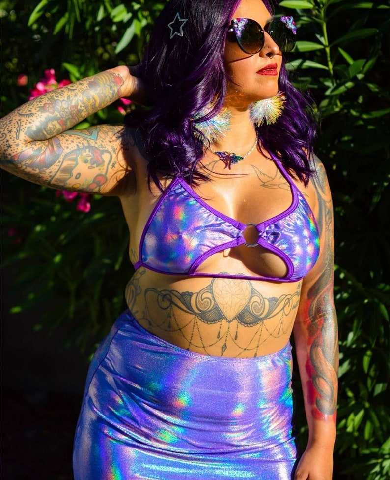 May include: A woman wearing a purple iridescent halter top with a ring detail and a matching skirt. She has purple hair, a star hair clip, and large feather earrings.