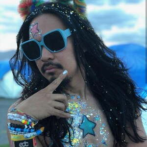 May include: A person with long black hair wearing blue square sunglasses and a rainbow fluffy headband. They have glitter on their chest and a blue star painted on their chest. They are wearing several bracelets on their wrist, including one that says "edc".