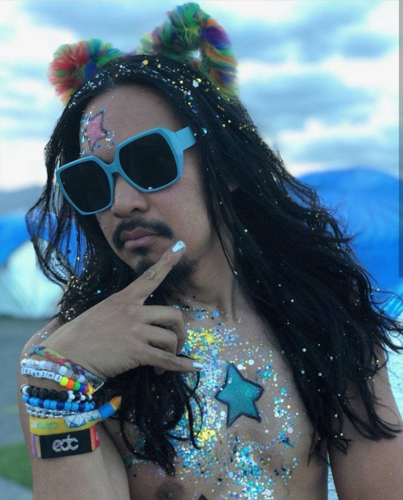 May include: A person with long black hair wearing blue square sunglasses and a rainbow fluffy headband. They have glitter on their chest and a blue star painted on their chest. They are wearing several bracelets on their wrist, including one that says "edc".