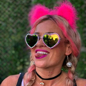 May include: A woman with pink hair is wearing heart-shaped sunglasses with iridescent rhinestones. She is also wearing a pink feather headband with fuzzy ear pieces and a black choker necklace with a rainbow charm.
