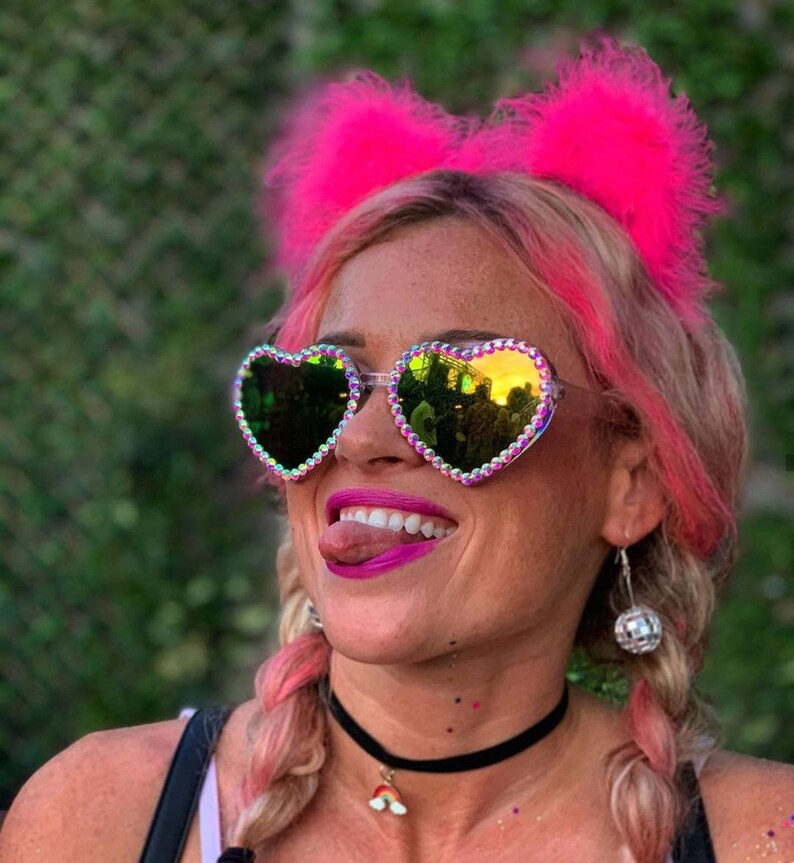 May include: A woman with pink hair is wearing heart-shaped sunglasses with iridescent rhinestones. She is also wearing a pink feather headband and a black choker necklace with a rainbow charm.