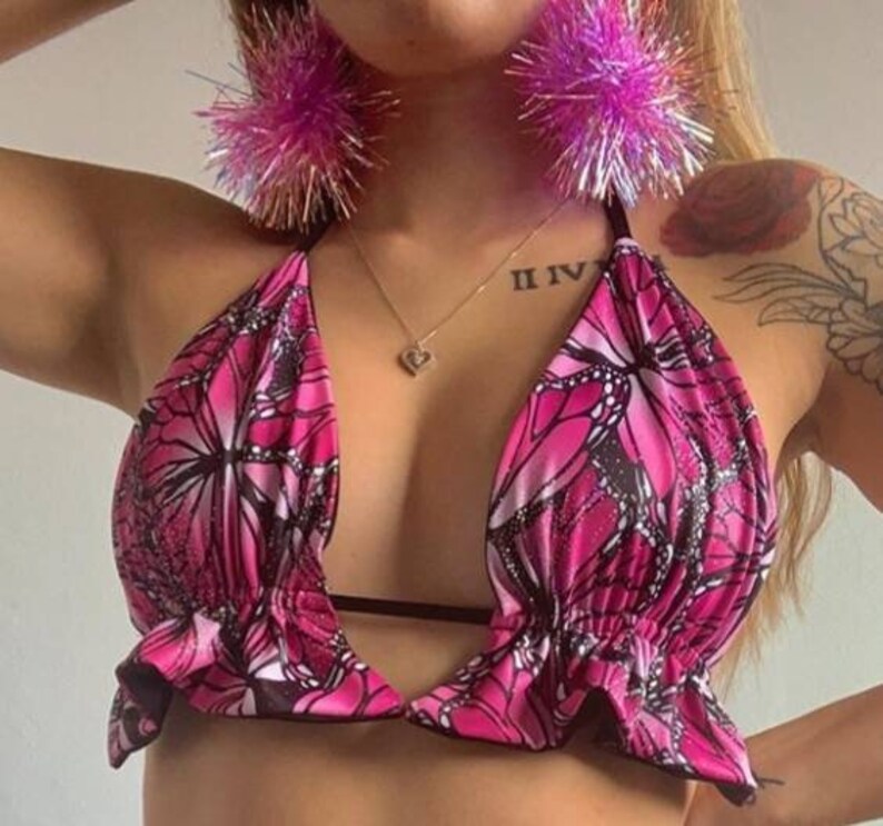 May include: Pink halter top with a butterfly print and ruffled trim. The top has a plunging neckline and a black strap across the chest.