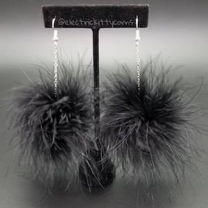 May include: A pair of black feather earrings with silver chains hanging from a black display stand. The earrings are made of fluffy black feathers and have a pom-pom shape.