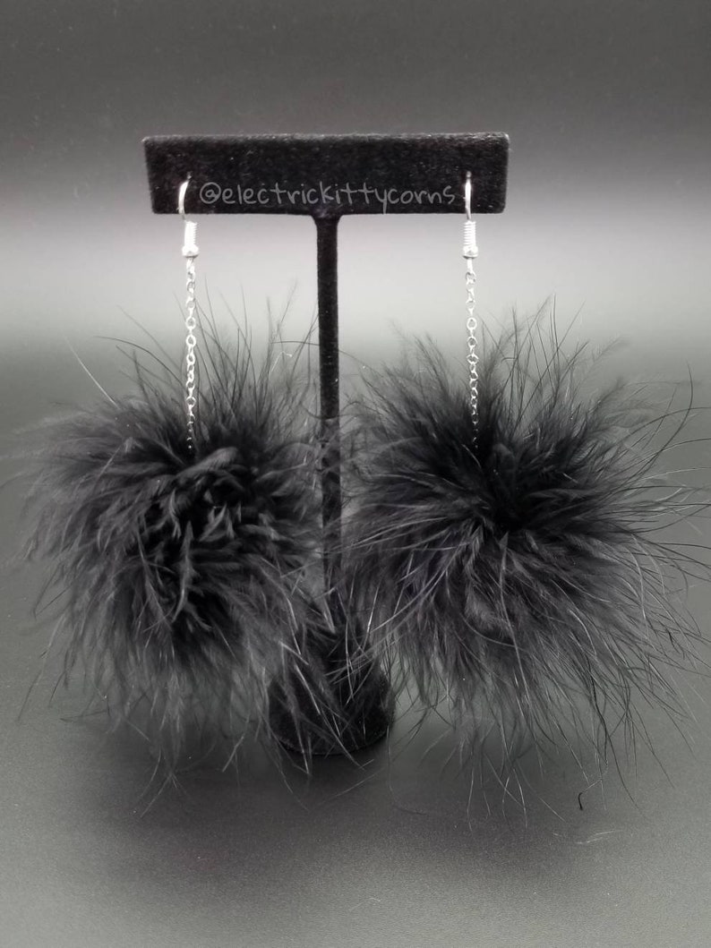 May include: A pair of black feather earrings with silver chains hanging from a black display stand. The earrings are made of fluffy black feathers and have a pom-pom shape.