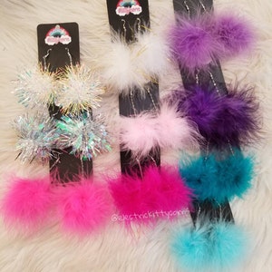 May include: Assortment of colorful fluffy and sparkly dangle earrings. The earrings are on display on black cards with the text "electric kittycorns" on one card.