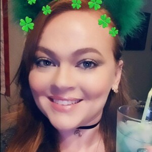May include: A woman with red hair is wearing a black choker necklace with a crescent moon charm. She is holding a tall glass of a green drink with ice and a straw. She is wearing green nail polish and has a green shamrock headband on her head.