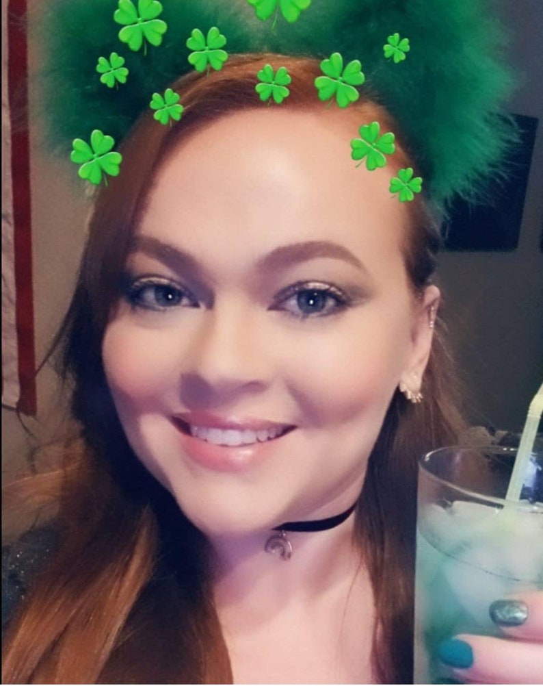 May include: A woman with red hair is wearing a black choker necklace with a crescent moon charm. She is holding a tall glass of a green drink with ice and a straw. She is wearing green nail polish and has a green shamrock headband on her head.