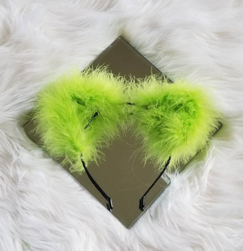 May include: A lime green feather headband with two large fluffy ear-like pieces. The headband is black and sits on a white fuzzy surface.