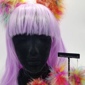 May include: A set of rainbow colored fluffy cat ears and earrings. The cat ears are on a purple wig and the earrings are on a black stand. The earrings are dangling and have a chain attached to them. The text '@electrickittycorns' is on the stand.