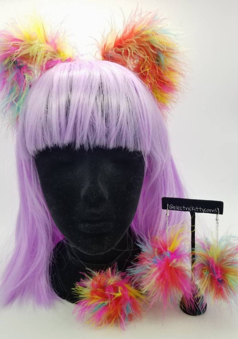 May include: A set of rainbow colored fluffy cat ears and earrings. The cat ears are on a purple wig and the earrings are on a black stand. The earrings are dangling and have a chain attached to them. The text '@electrickittycorns' is on the stand.