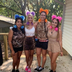 May include: Four women wearing leopard print outfits and fuzzy animal ear headbands. The headbands are purple, blue, orange, and pink. The women are standing on a patio with a wooden railing in the background.