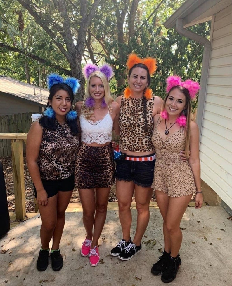 May include: Four women wearing leopard print outfits and fuzzy animal ear headbands. The headbands are purple, blue, orange, and pink. The women are standing on a patio with a wooden railing in the background.