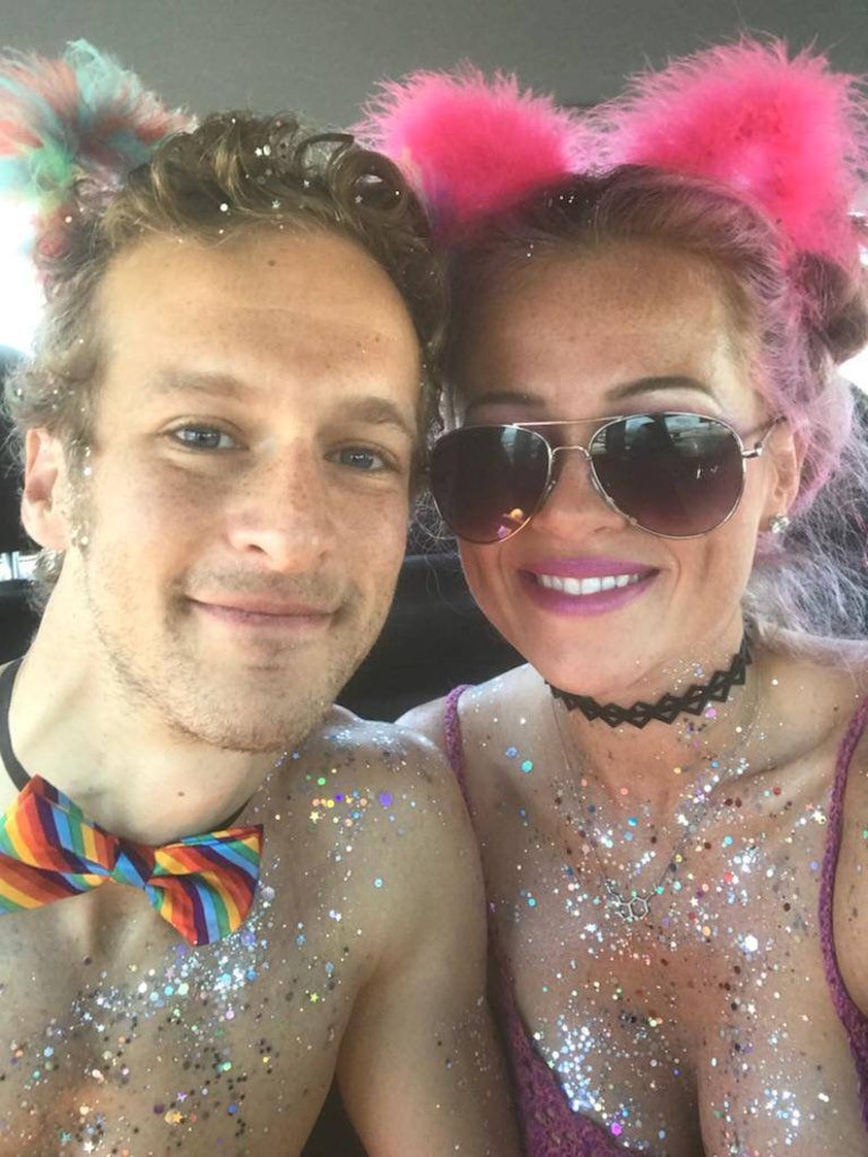 May include: A couple covered in glitter, wearing festive accessories. The woman has pink hair and is wearing a pink furry headband with cat ears. The man is wearing a rainbow bow tie.