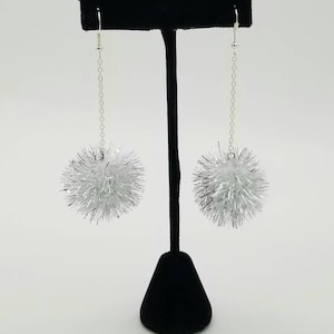 Pode incluir: Um par de brincos de prata com detalhes de pompom. Os brincos estão pendurados em um suporte de brincos preto.