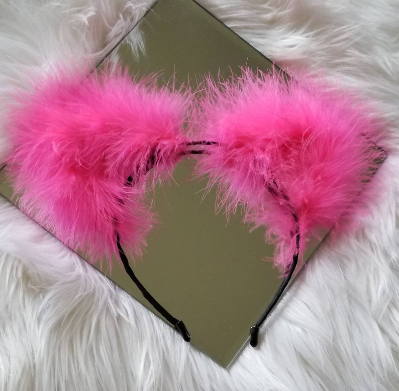May include: A pink fluffy headband with two large, feathery ears. The headband is black and has a simple design.