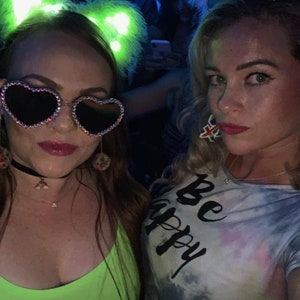 May include: Two women wearing colorful outfits and sunglasses pose for a photo. One woman is wearing a green top and a headband with glowing cat ears. The other woman is wearing a tie-dye shirt with the text "Be Happy" and a white headband with sparkly feather accents.