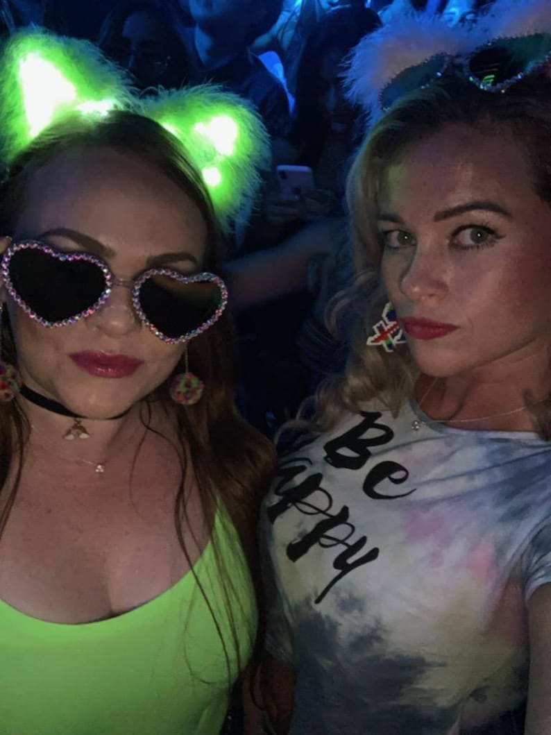 May include: Two women wearing colorful outfits and sunglasses pose for a photo. One woman is wearing a green top and a headband with glowing cat ears. The other woman is wearing a tie-dye shirt with the text "Be Happy" and a white headband with sparkly feather accents.