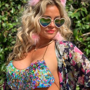 May include: A woman wearing a colorful iridescent sequined bra top with a chain strap. She is also wearing heart-shaped sunglasses with rhinestones and pink fuzzy earmuffs.