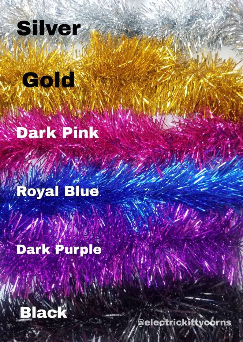 May include: A close-up of six spools of metallic tinsel garland. The spools are arranged vertically, with the colors silver, gold, dark pink, royal blue, dark purple, and black. The text "@electrickittycorns" is visible at the bottom of the image.
