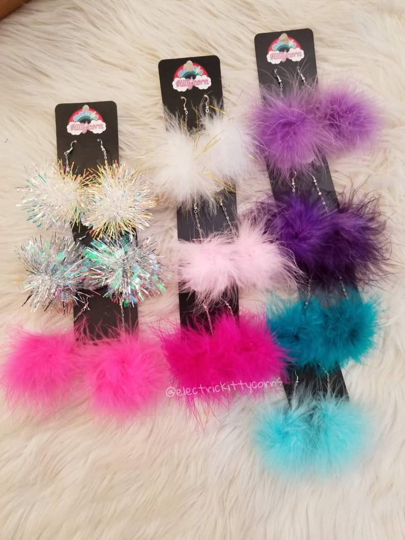 May include: Assortment of colorful fluffy and sparkly dangle earrings. The earrings are on display on black cards with the text "electric kittycorns" on one card.