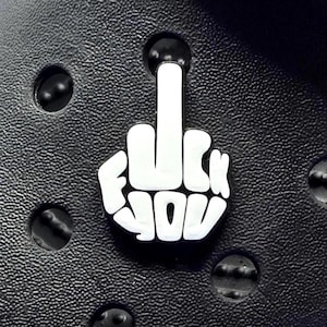 May include: White shoe charm in the shape of a hand giving the middle finger, with the words "FUCK YOU" in a bold font. The charm is set against a black background with circular holes.