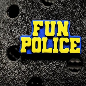 May include: A yellow and blue shoe charm with the words "FUN POLICE" in bold, block letters. The charm is set against a textured black surface with circular holes, likely a shoe. The text is yellow with a blue outline.