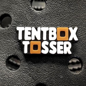 May include: A black shoe charm featuring the text "TENTBOX TOSSER" in white with orange square details. The charm is set on a textured black surface with circular perforations.
