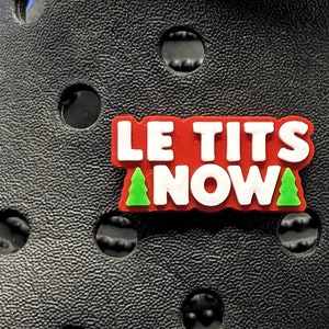 May include: Close-up of a black clog shoe with a decorative charm. The charm has the words "LE TITS NOW" in white letters on a red background, with green Christmas trees. The shoe has multiple round holes.