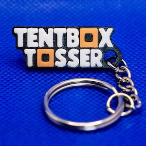 May include: A black and white keyring with the text "TENTBOX TOSSER" in white letters with orange accents. The keyring has a silver chain and ring, set against a blue background.