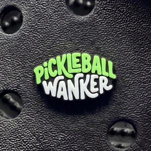May include: A black shoe charm featuring the text PICKLEBALL WANKER in lime green and white lettering. The charm is circular with a textured surface and is inserted into a black foam clog with multiple circular ventilation holes visible.
