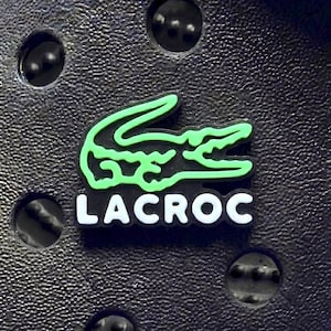 May include: A black textured foam clog accessory featuring a bright green crocodile graphic positioned above the white text LACROC. The emblem is centred within a circular ventilation hole pattern on the surface of the black footwear material.