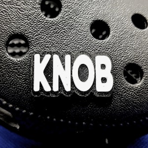 May include: Close-up of a black shoe with the word "KNOB" in white block letters. The shoe has several round holes and a textured surface. The word "KNOB" is in a rectangular shape.