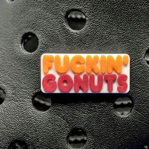 May include: A white rectangular shoe charm with the words "FUCKIN' GONUTS" in orange and red lettering. The charm is set against a textured black surface with circular holes.