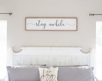 May include: White wooden headboard with a white and black striped pillow and a white pillow with black cursive text that says "Let's Get Cozy". Above the headboard is a wooden sign with the text "stay awhile" in black cursive.