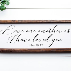 May include: A wooden framed sign with a white background and black text. The text reads "Love one another as I have loved you" and "John 15:12".