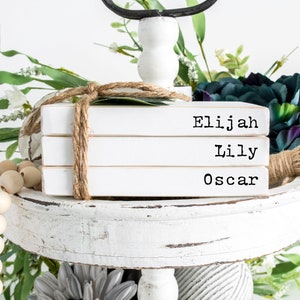 May include: Three white books stacked on a tiered tray. The books are tied together with twine and have the names "Elijah", "Lily", and "Oscar" printed on them in black.