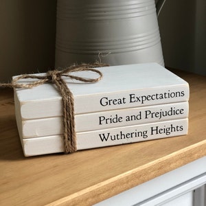 May include: Three white painted books tied with twine, featuring the titles "Great Expectations", "Pride and Prejudice", and "Wuthering Heights" in black. The books are displayed on a wooden surface.