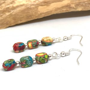 May include: Dangle earrings with rectangular beads in red, blue, green, and yellow. The beads are strung on silver-coloured wire with a hook closure. The earrings are displayed against a white background. Each earring has three beads.