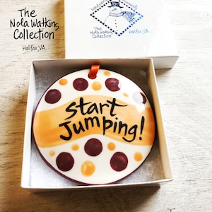 May include: A round, white ceramic ornament with brown and orange polka dots and a yellow stripe. The ornament says "Start Jumping!" in black cursive writing.