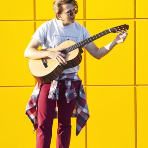 Może przedstawiać: Młoda osoba w szarej koszulce z napisem "CH", czerwonych spodniach i kratkowanej koszuli zawiązanej w talii, gra na brązowym akustycznym gitarze przed ż&oacute;łtą ścianą.
