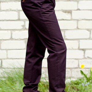 May include: A pair of dark purple pants with a brown leather patch on the back pocket. The pants are worn by a person standing in front of a white brick wall.