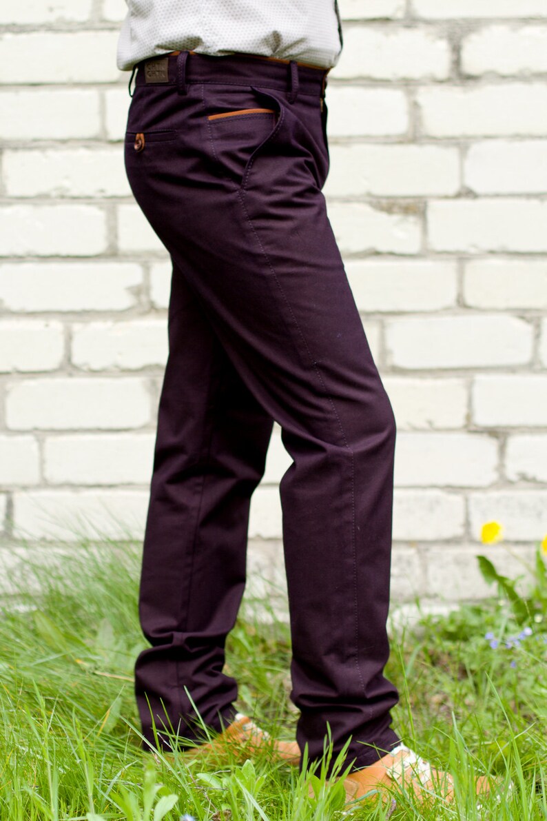May include: A pair of dark purple pants with a brown leather patch on the back pocket. The pants are worn by a person standing in front of a white brick wall.