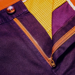 May include: Close-up of a purple pair of pants with a gold zipper and a brown button. The zipper is partially open, revealing a yellow and white checkered lining.