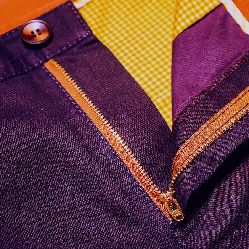 May include: Close-up of a purple pair of pants with a gold zipper and a brown button. The zipper is partially open, revealing a yellow and white checkered lining.