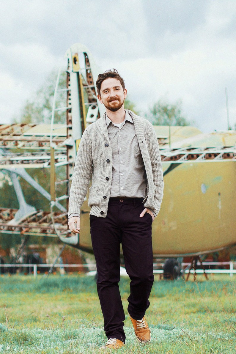 May include: A man wearing a light brown cardigan over a light gray button-down shirt and dark blue pants. He is standing in front of a large metal structure.